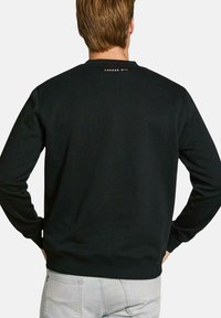 Sudadera negra de algodón, con mangas largas y cuello redondo. El texto "LONDON W '11" está impreso en blanco en la parte superior de la espalda.