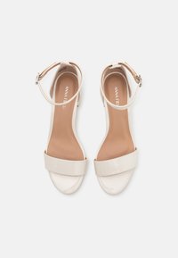 Beige patent lærred hæl sandaler med rund tå, ankelrem og bred frontbånd. Glat tekstur med minimalistisk design.