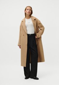 Lungo cappotto beige con vestibilità rilassata, due bottoni e ampi rever, abbinato a una maglia bianca e pantaloni neri a vita alta; texture liscia.