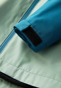 Ärmel einer türkisfarbenen Jacke mit Klettverschluss auf hellgrünem Stoffhintergrund. Glatte Textur mit sauberer Verarbeitung entlang der Nähte.