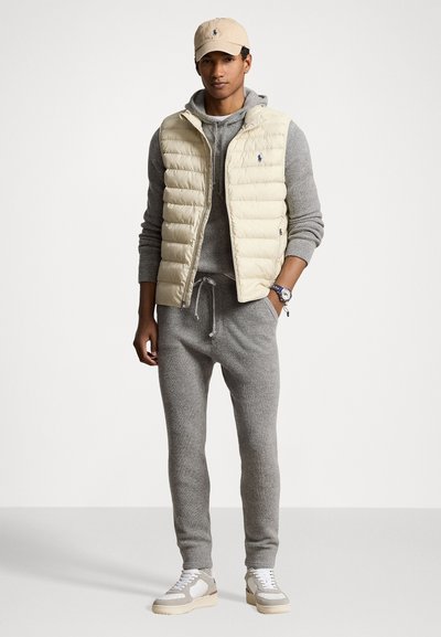 Gilet matelassé beige sur un sweat à capuche en tricot gris et un pantalon de survêtement assorti. Des baskets blanches complètent la tenue. Logo discret sur le gilet et le bonnet.
