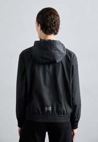 Jaqueta leve preta com capuz, punhos elásticos e barragem. Apresenta um discreto logotipo dourado nas costas. Superfície suave e design minimalista.
