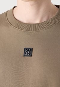 Gros plan d'un t-shirt beige à col rond avec un petit patch logo carré noir indiquant "HUGO" sur la poitrine.