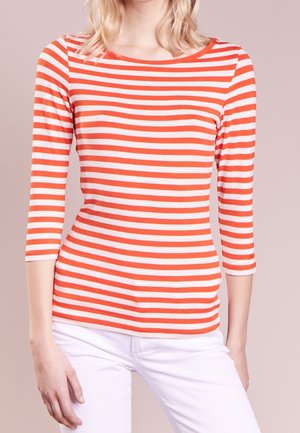 Personne portant un t-shirt ajusté à manches longues rayé horizontalement orange et blanc, assorti à un pantalon blanc, sur un fond uni.