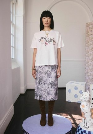 Kvinde står på en rund platform iført en hvid t-shirt med grafik, et mønstret lavendelskjørt, brune støvler og lag på lag halskæder, i et lyst rum med katte-skulpturer.