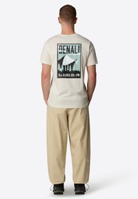 Creamfarvet bomulds-t-shirt med et Denali grafisk tryk på ryggen. Kombineret med løse beige bukser og sorte sko, der viser en afslappet pasform.