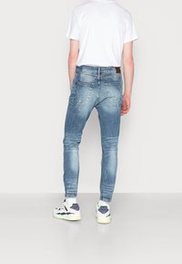 Slim-fit modré džínové kalhoty s vybledlým povrchem, zadními kapsami a hnědou koženou štítkem na pase, doplněné bílými a vícebarevnými teniskami.