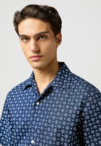 Camisa de botón azul marino con un patrón floral, que presenta flores en un contraste de azul claro, cuello y mangas cortas. Tejido de algodón.