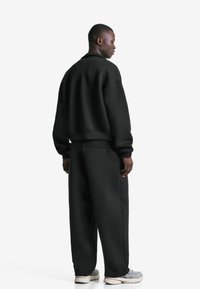 Mand, der står vendt til siden iført en sort oversized jakke og matchende løstsiddende bukser med grå og hvide sneakers på en hvid baggrund.