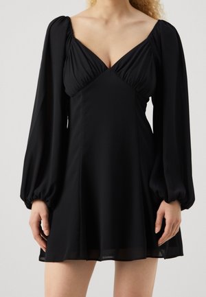 Vrouw in een zwarte lange mouwen mini-jurk met een diepe V-hals en plooidetails op de buste en mouwen.