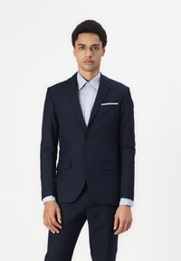 Navy suit jacket with two buttons, notched lapels, and front pockets. Light blue shirt underneath with a pointed collar and subtle pocket detail.