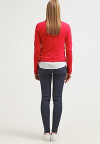 Pull rouge à manches longues porté sur une chemise blanche, associé à un jean skinny bleu foncé et des baskets blanches, vu de dos.
