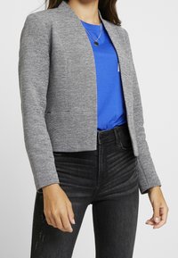 Veste grise texturée avec un design raccourci, une fermeture à un seul bouton et un col arrondi, superposée à un t-shirt bleu et un jean noir.