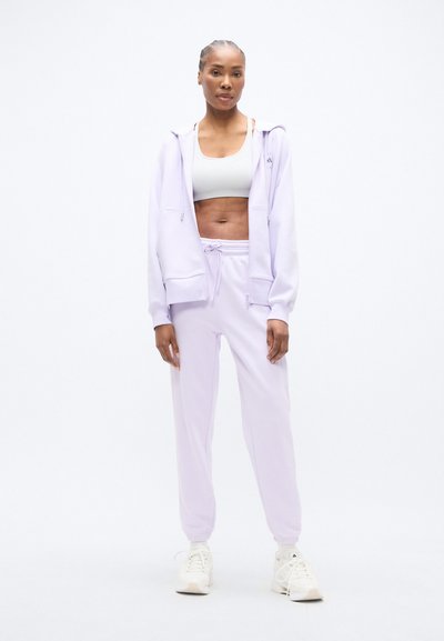 Svetlofialový športový set pozostávajúci z mikiny na zips s kapucňou a nohavíc so sťahovacou šnúrkou, pod ktorým je sivý crop top. Hladká textúra a voľný strih.