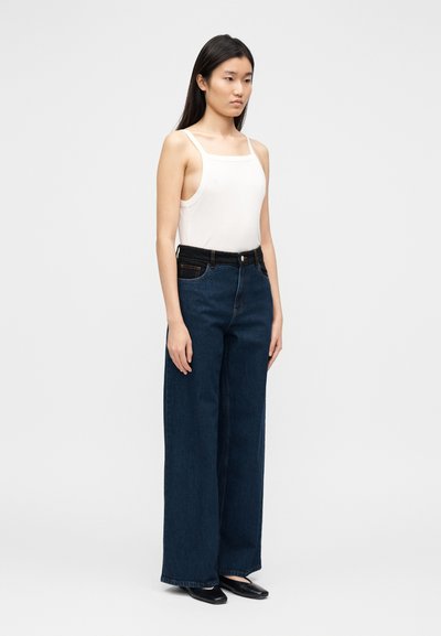 Λευκό αμάνικο τοπ και σκούρο μπλε τζιν παντελόνι με φαρδιά μπατζάκια και μαύρη ζώνη στη μέση. Ομαλή υφή, διακριτικές ραφές και ελάχιστα μεταλλικά στοιχεία.