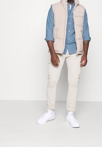 Gilet imbottito beige sopra una camicia azzurro chiaro con bottoni, abbinato a pantaloni cargo beige e scarpe da ginnastica bianche. Dotato di cerniere e tasche.