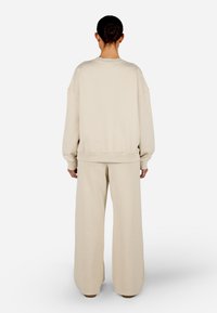 Bēšas pārāk liela izmēra sporta krekls ar ribotām aprocēm un apakšu, kombinācijā ar atbilstošiem brīvi pieguļošiem biksēm. Mīksta, teksturēta auduma. Aizmugures skats.