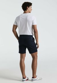 Weißes T-Shirt mit kurzen Ärmeln, marineblaue Shorts und weiße Turnschuhe mit roten Akzenten. Freizeitkleidung, die eine figurbetonte Silhouette und ein sauberes Design präsentiert.