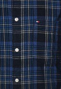 Sininen ja musta ruudullinen napillinen paita, jossa on rintatasku. Paidassa on valkoiset napit ja erilaisissa sävyissä olevia pystysuoria raitoja.