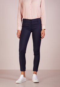 Pantalones ajustados azul marino con costuras frontales, combinados con una camisa de botones rosa claro y zapatillas blancas, sobre un fondo neutro.
