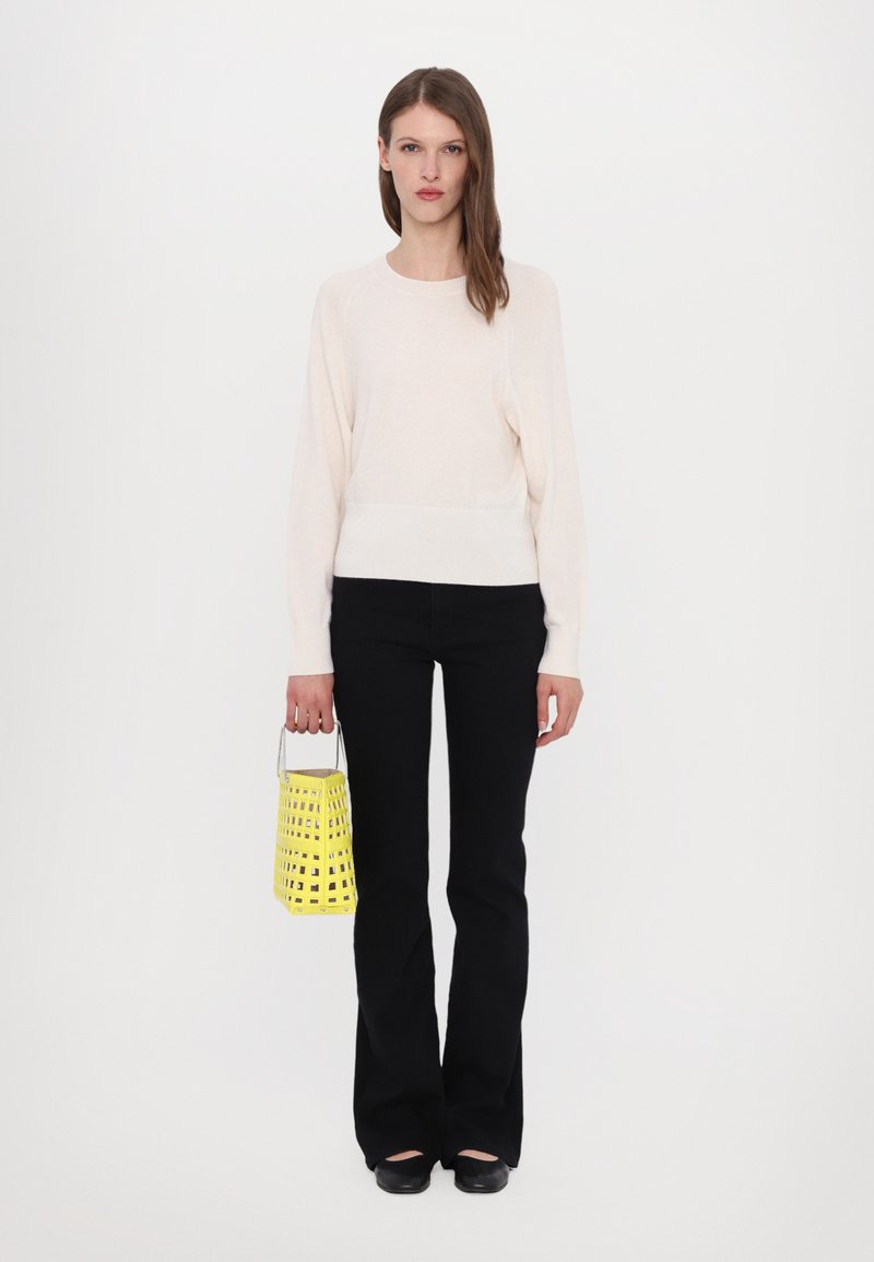 Cremefarbener Strickpullover, schwarze Schlaghosen und eine kleine gelbe Bucket Bag mit Ausschnittmuster. Das Schuhwerk ist schwarz mit runder Zehenpartie.