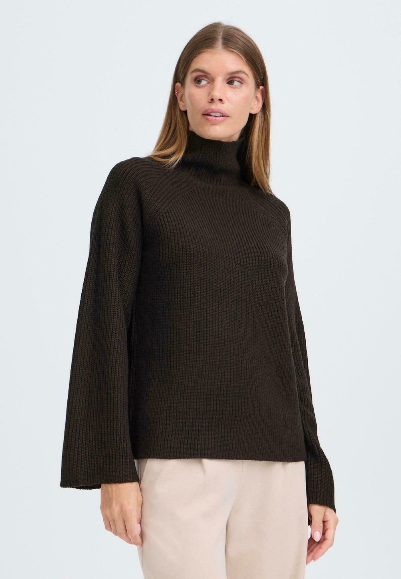 Dunkelbrauner, gerippter Strickpullover mit hohem Kragen und weiten Ärmeln, getragen über hellbeigen Hosen. Die Textur wirkt weich und gemütlich.