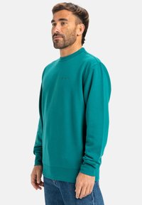 Teal sweatshirt med rund hals, lange ærmer og ribbede manchetter. Har subtile logo detaljer i matchende farve på brystet.