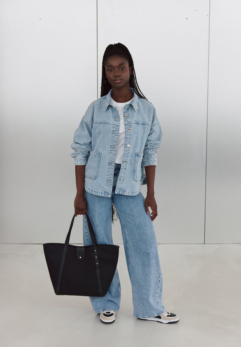 Veste en denim avec grandes poches, couleur bleu clair, associée à un jean bleu clair ample. Tenant un sac fourre-tout noir, sneakers noir et blanc.