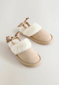 Beige čībiņas ar pūkainu baltu apkakli, mīkstu zamšādas virsu un elastīgu lentu ar odziņu. Nedaudz kontūrēta gumijas zole.