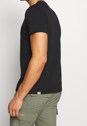 Homme portant un t-shirt noir à manches courtes et un pantalon cargo vert Columbia, debout de profil sur un fond blanc.