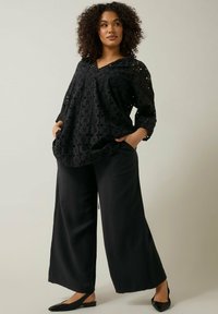 Schwarze Bluse mit Lochmuster, V-Ausschnitt und Dreiviertelärmeln. Kombiniert mit lockeren schwarzen weit geschnittenen Hosen und schwarzen flachen Schuhen.