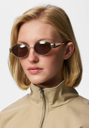 Kvinde med blond bob-frisure iført ovale brune solbriller og beige jakke med lynlåst høj krave.