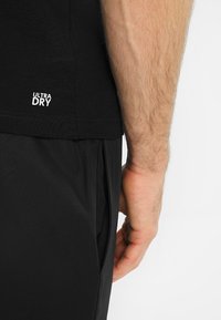 Maglietta atletica nera con logo "ULTRA DRY", maniche corte e tessuto testurizzato, abbinata a pantaloni neri. In primo piano spalle e braccio.