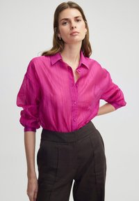 Fuchsiová průhledná košile na knoflíky s límečkem a vyhrnutými rukávy. V kombinaci s tmavě hnědými kalhotami na míru, které zvýrazňují kontrastní barvy.
