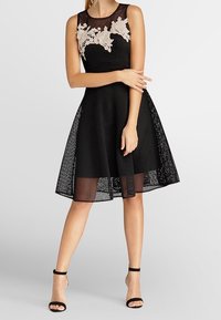 Robe A-line sans manches en mesh noir avec des appliqués floraux beiges au niveau du corsage, portée avec des escarpins ouverts à bout découpé et à bride à la cheville noires.