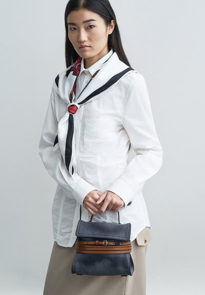 Mulher com cabelo longo e escuro, vestindo uma camisa branca de botões, um lenço estampado, uma saia bege e segurando uma mala cinza com detalhes em cinto castanho.