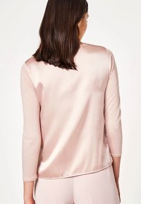 Ljusrosa satinblus med en mjuk textur, långa ärmar och rundad nederkant, sedd bakifrån. Minimalistisk design, utan utsmyckningar.