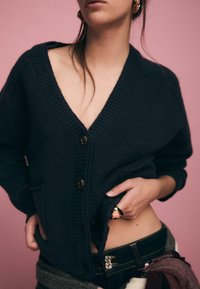 Dunkelblauer Cardigan aus weichem Stoff mit V-Ausschnitt, gerippten Abschlüssen und vier großen Knöpfen, kombiniert mit einer Jeans in der Taille.