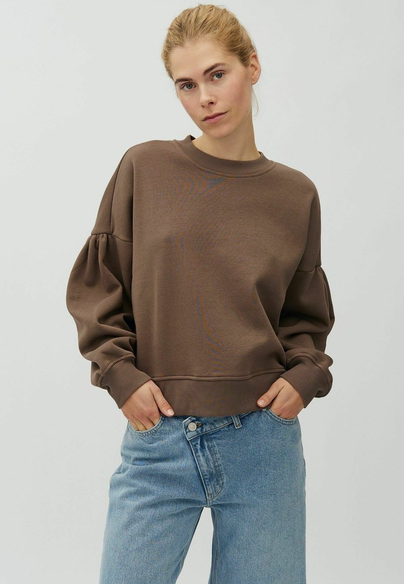 Femme aux cheveux blonds portant un pull marron à manches bouffantes et un jean bleu clair, debout devant un fond clair uni, les mains dans les poches.