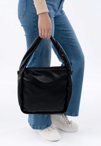 Musta nahkalaukku pehmeällä pinnalla, vetskarisuljenta ja kaarevat, pehmustetut kahvat, jota pitää henkilö, joka on pukeutunut sinisiin farkkuihin ja valkoisiin lenkkitossuihin.