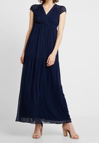 Žena oblečená v námořnické modré maxi šatech s krátkým rukávem a výstřihem do V, v tělových sandálech na vysokém podpatku, stojí na neutrálním pozadí.