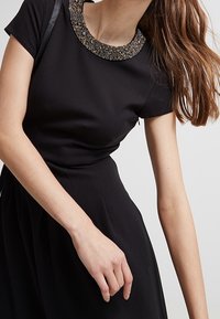 Schwarzes Kleid mit taillierter Oberpartie und schwingendem Rock. Verfügt über einen strukturierten Stoff und einen mit Schmucksteinen besetzten Kragen mit metallischen Akzenten. Kurzärmlig.