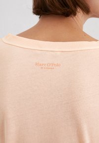 Helles Pfirsichfarbenes T-Shirt mit kurzen Ärmeln aus weichem Material. Ausgestattet mit einem gerippten Halsausschnitt und einem geprägten Markenetikett auf der Rückseite.