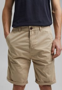 Beżowe bawełniane szorty z podwiniętym dołem, z przodem zapinanym na guzik, bocznymi kieszeniami i gładką teksturą. W połączeniu z ciemną koszulą.