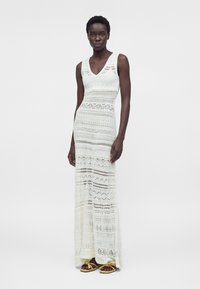 Femme portant une robe maxi blanche en crochet sans manches et des sandales jaunes, debout devant un fond clair uni.