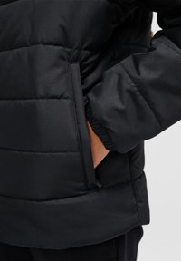 Sort puffjakke med quiltet tekstur, der har en sidelomme og elastiske manchetter, lavet af glat, let væv.