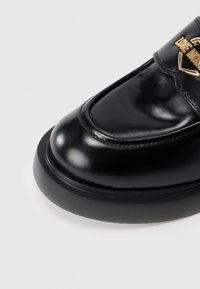 Sapato de couro preto com um acabamento brilhante, apresentando bico arredondado, sola grossa preta e um enfeite em forma de coração dourado na tira.
