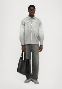 Cămașă din denim gri cu nasturi, având o buzunar pe piept, asortată cu blugi gri închis. Modelul ține o geantă de mână din piele neagră cu o textură netedă.
