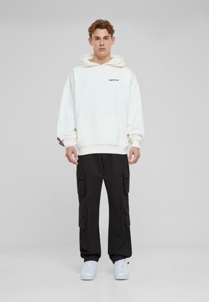 Hvit hettegenser med frontlomme og svart logo, sammen med svarte cargo-bukser med flere lommer, og hvite sneakers.