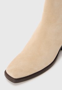 Stivale alla caviglia in suede beige con punta quadrata, caratterizzato da una suola marrone scuro e dettagli di cucitura lungo il bordo. Texture liscia.
